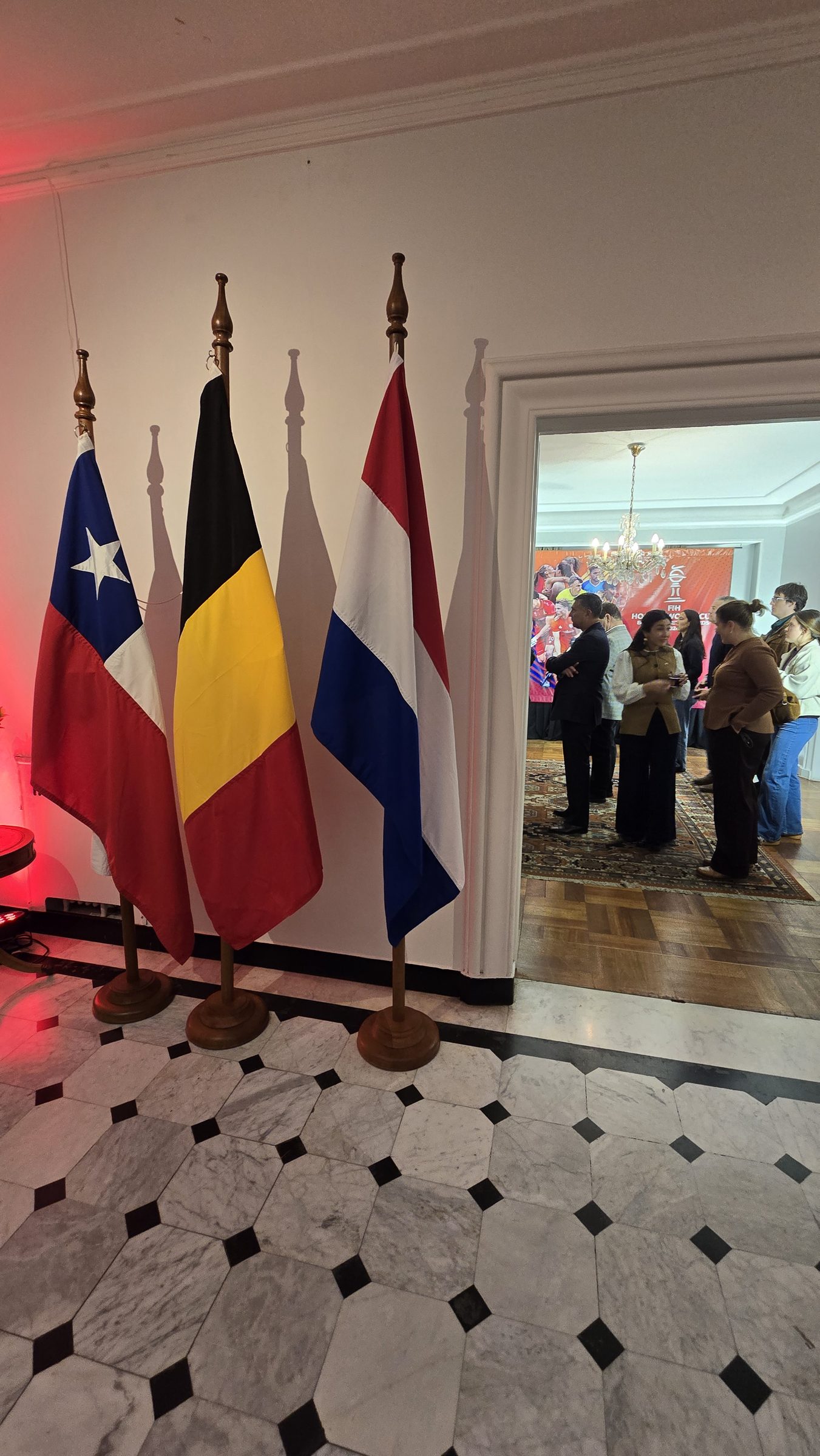 Cuatro banderas sobre pies de madera en una sala de recepción, con gente reflejada en un espejo al fondo