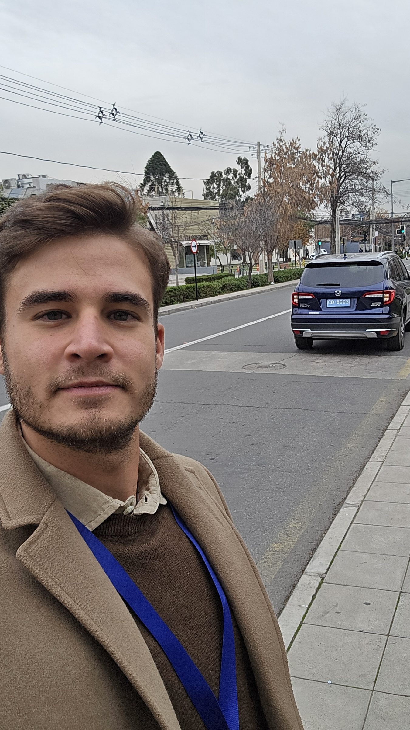 Selfie en una calle de Santiago con una acreditación al cuello; al fondo, un coche con matrícula diplomática