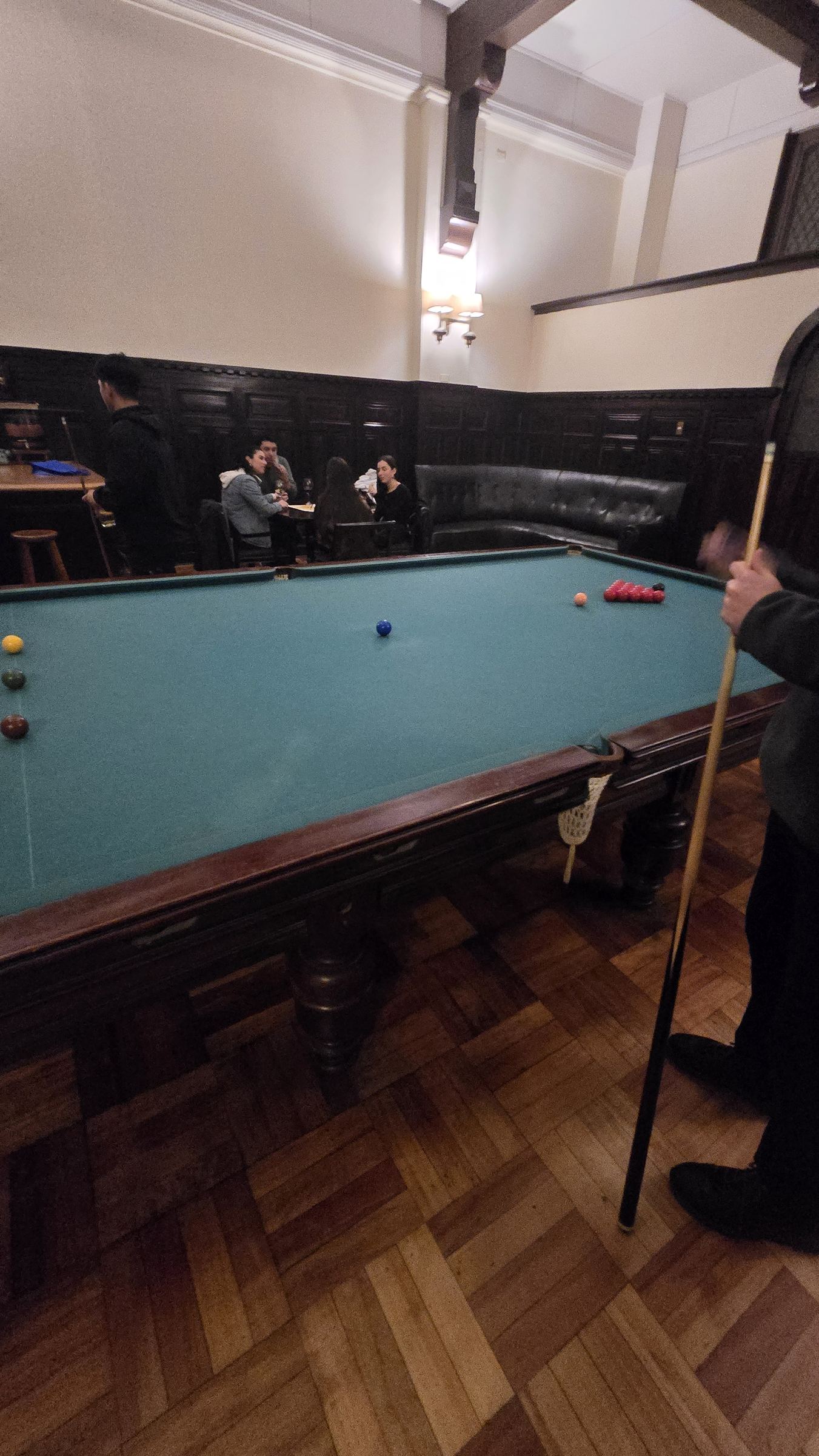 Una partida de billar en un salón antiguo, con más gente alrededor de una mesa al fondo