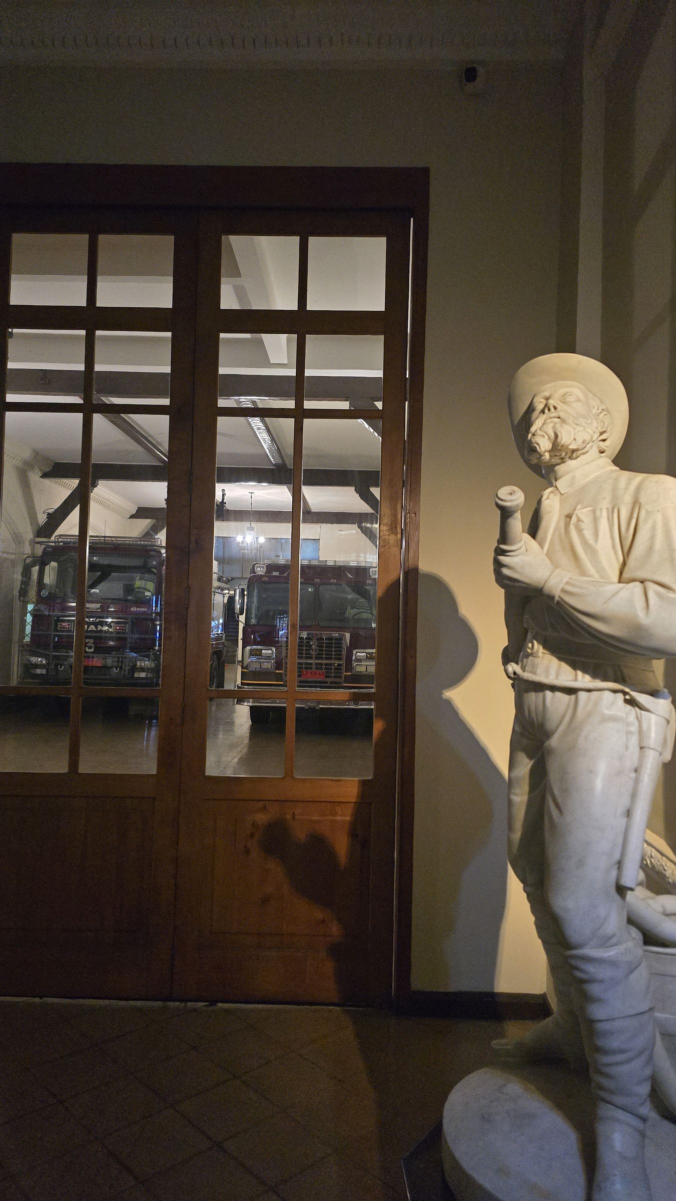Una estatua de bombero junto a la entrada de un cuartel, con los carros vistos a través de una puerta de cristal al fondo
