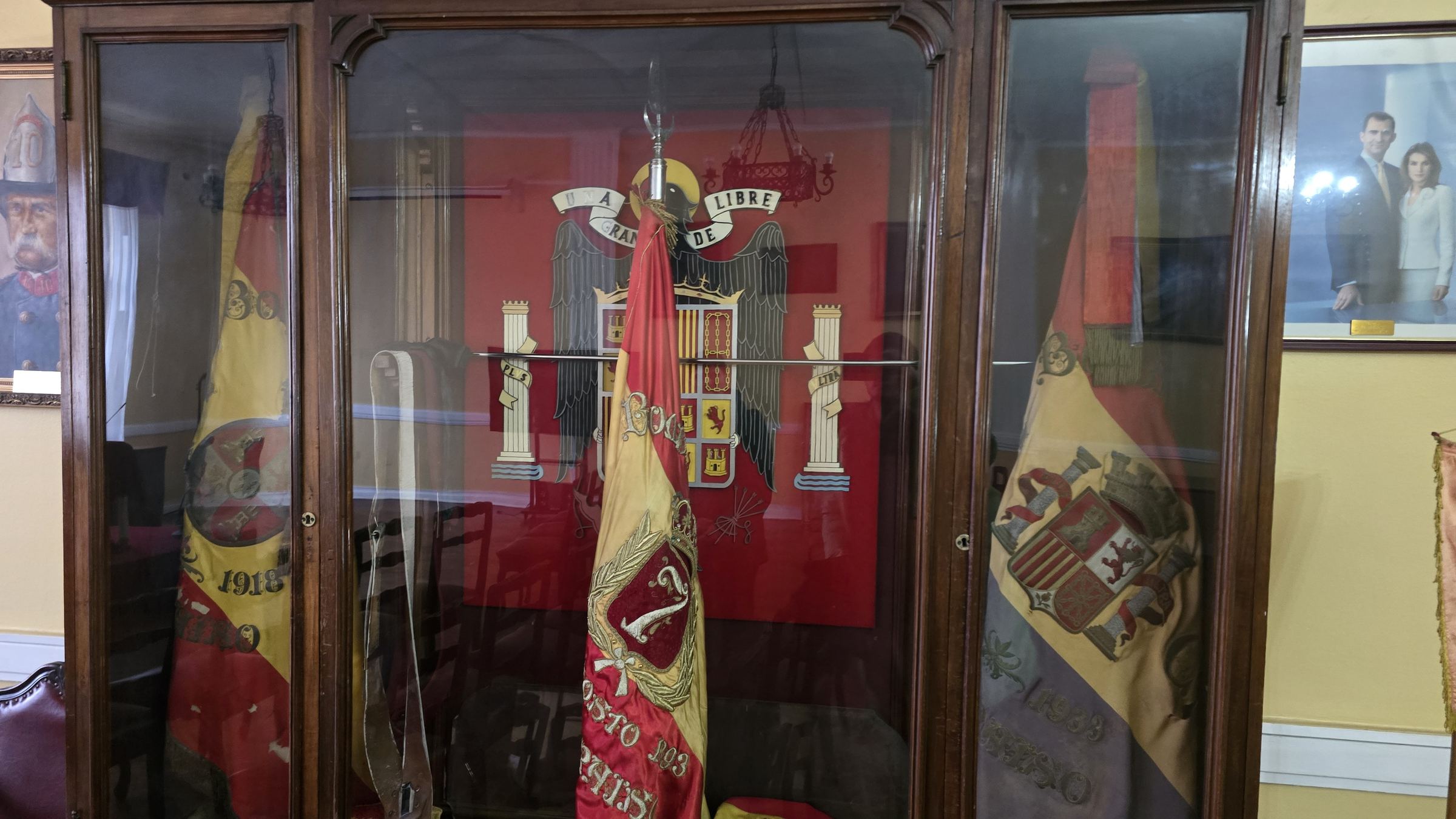 Una vitrina con banderines y estandartes históricos españoles, y un retrato de los reyes de España