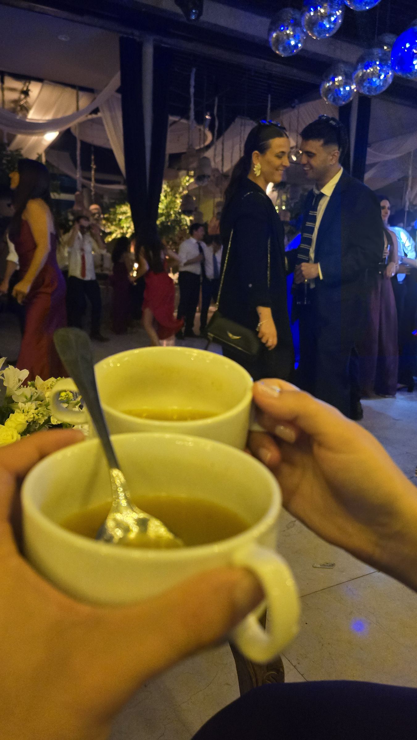 Dos manos sosteniendo tazas de consomé, con una pareja conversando al fondo
