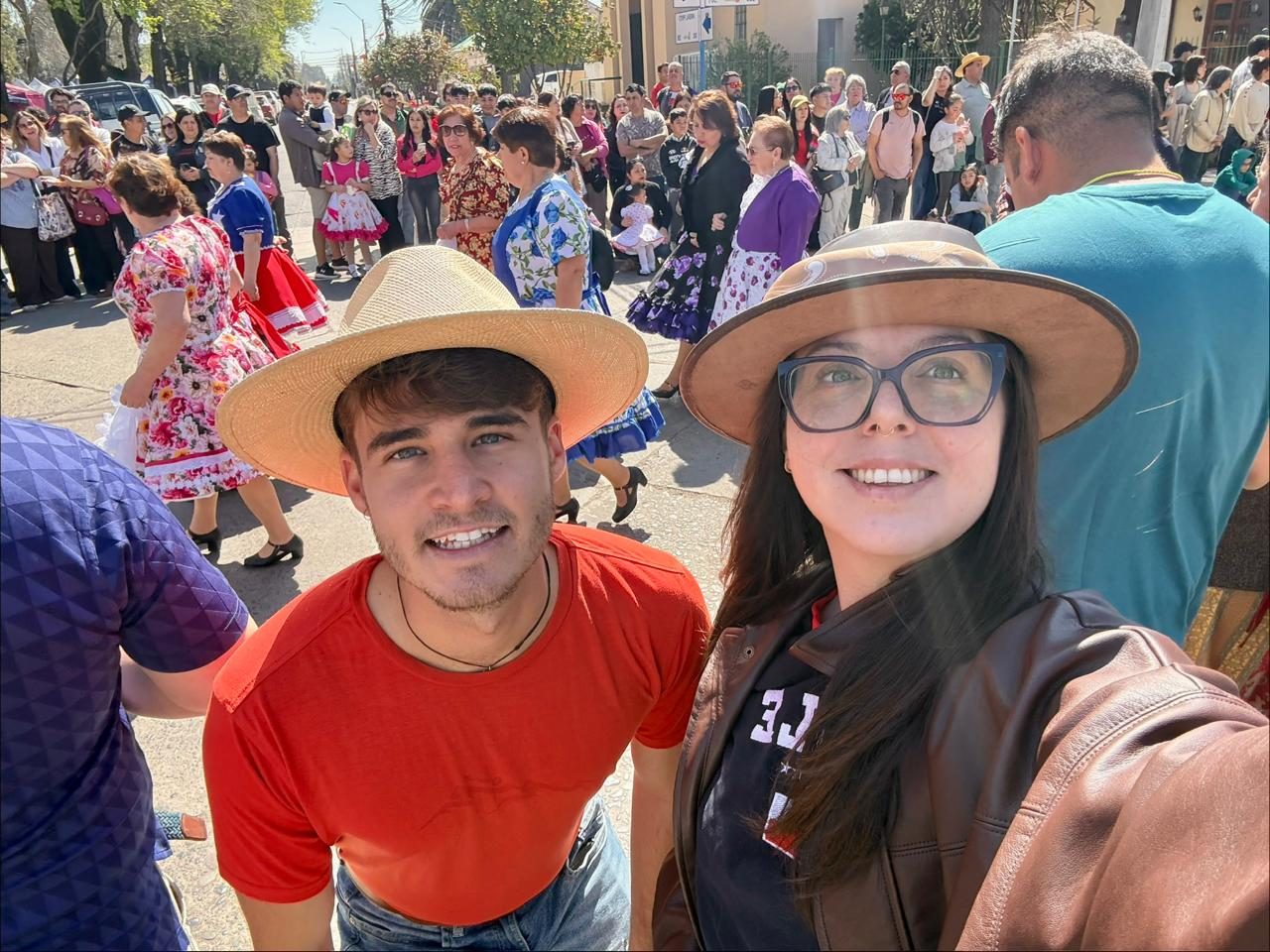 Un hombre y una mujer con sombreros de huaso se fotografían en la calle, con parejas bailando cueca y público al fondo