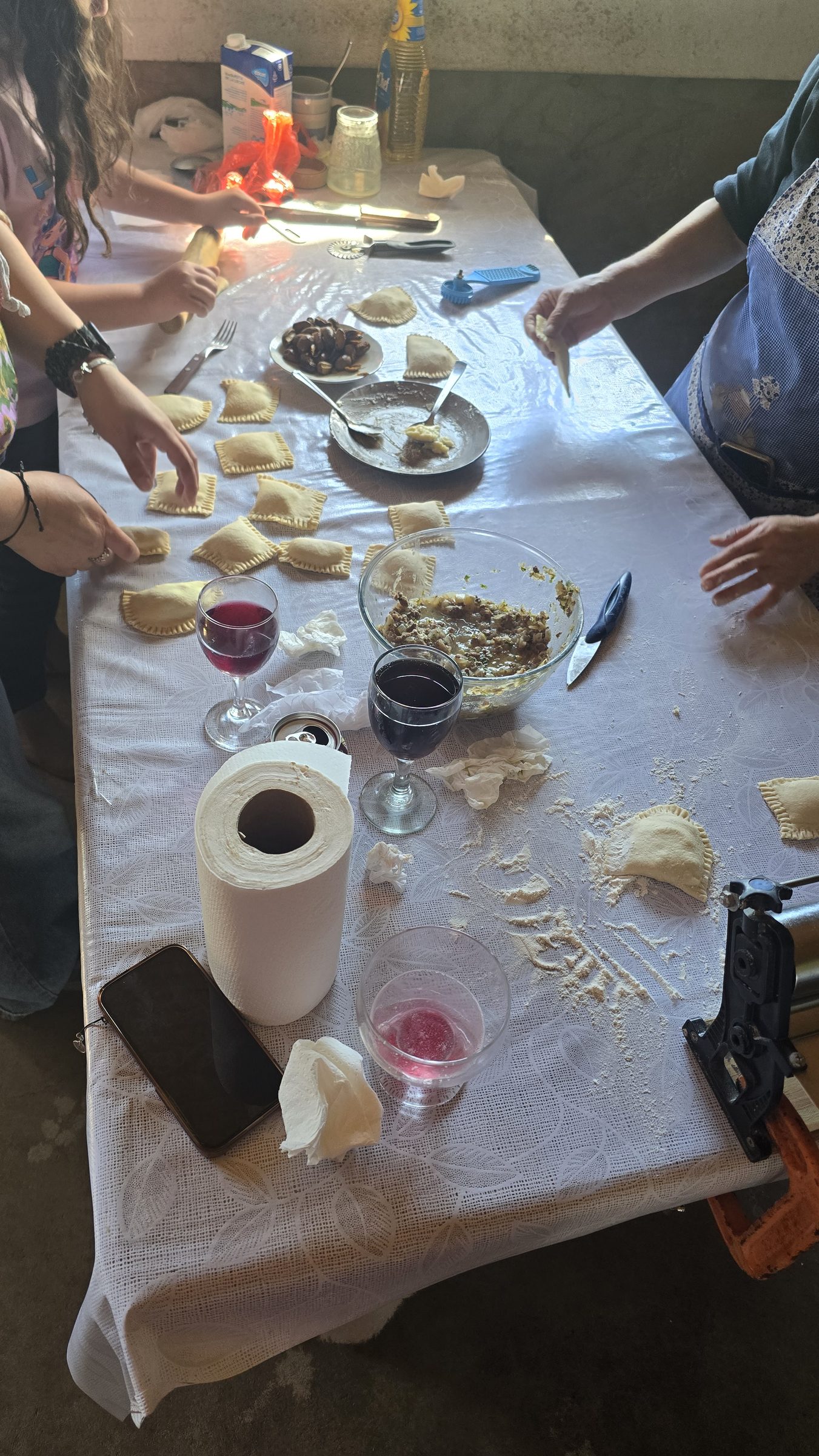 Varias personas arman empanadas sobre una mesa cubierta con mantel, con copas de vino y un bol de pino cerca
