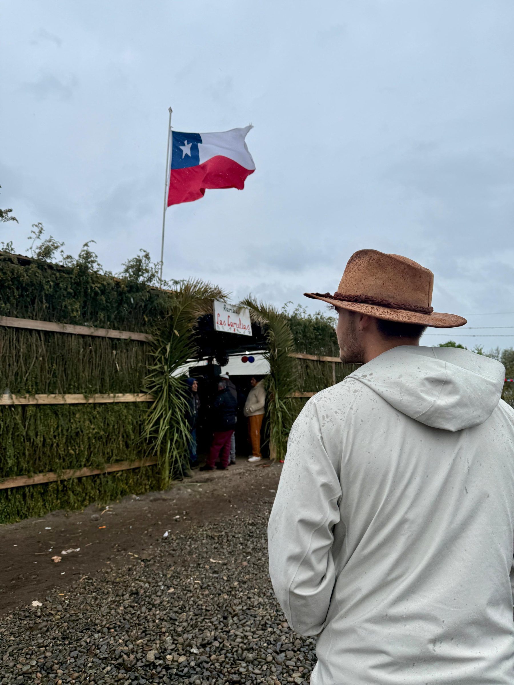 Un hombre con sombrero mira hacia la entrada de una fonda bajo la lluvia, con una bandera chilena ondeando en lo alto