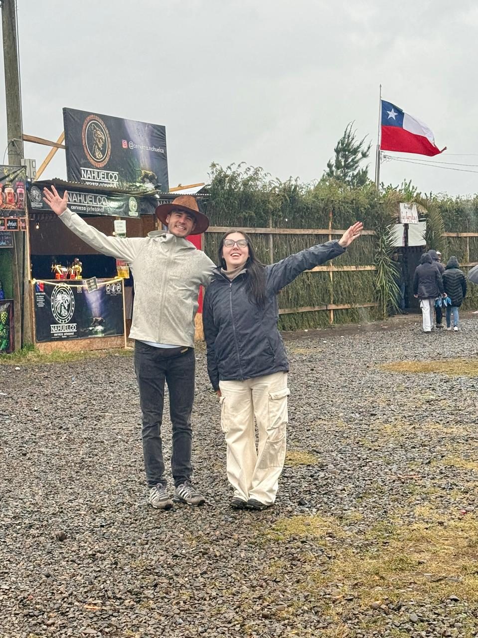 Una pareja con los brazos en alto celebra bajo la lluvia frente a la entrada de una fonda, con una bandera chilena ondeando al fondo