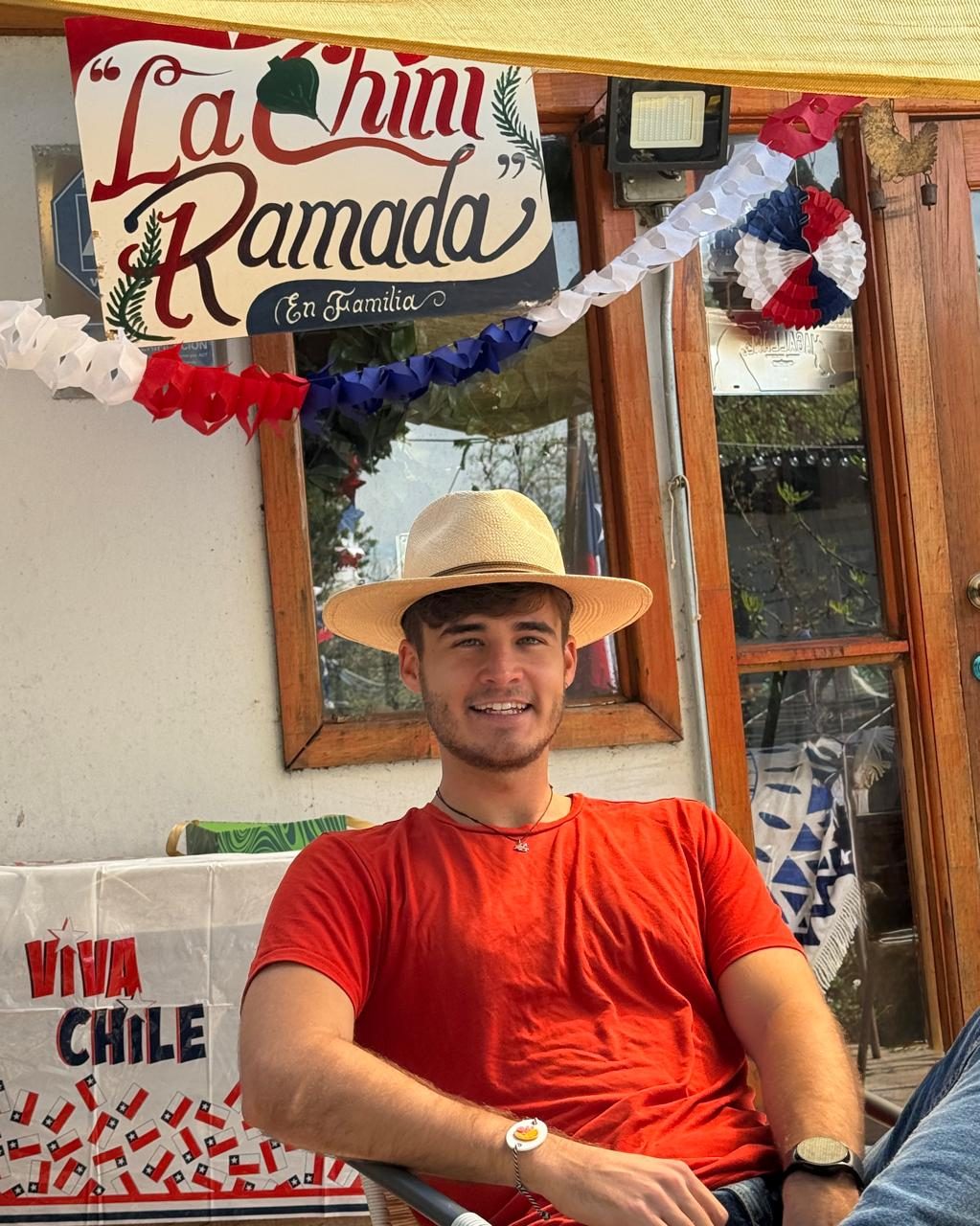 Un joven con sombrero y camisa roja sonríe sentado bajo un cartel que dice «La Chini Ramada», decorado con banderines rojos, blancos y azules de Fiestas Patrias